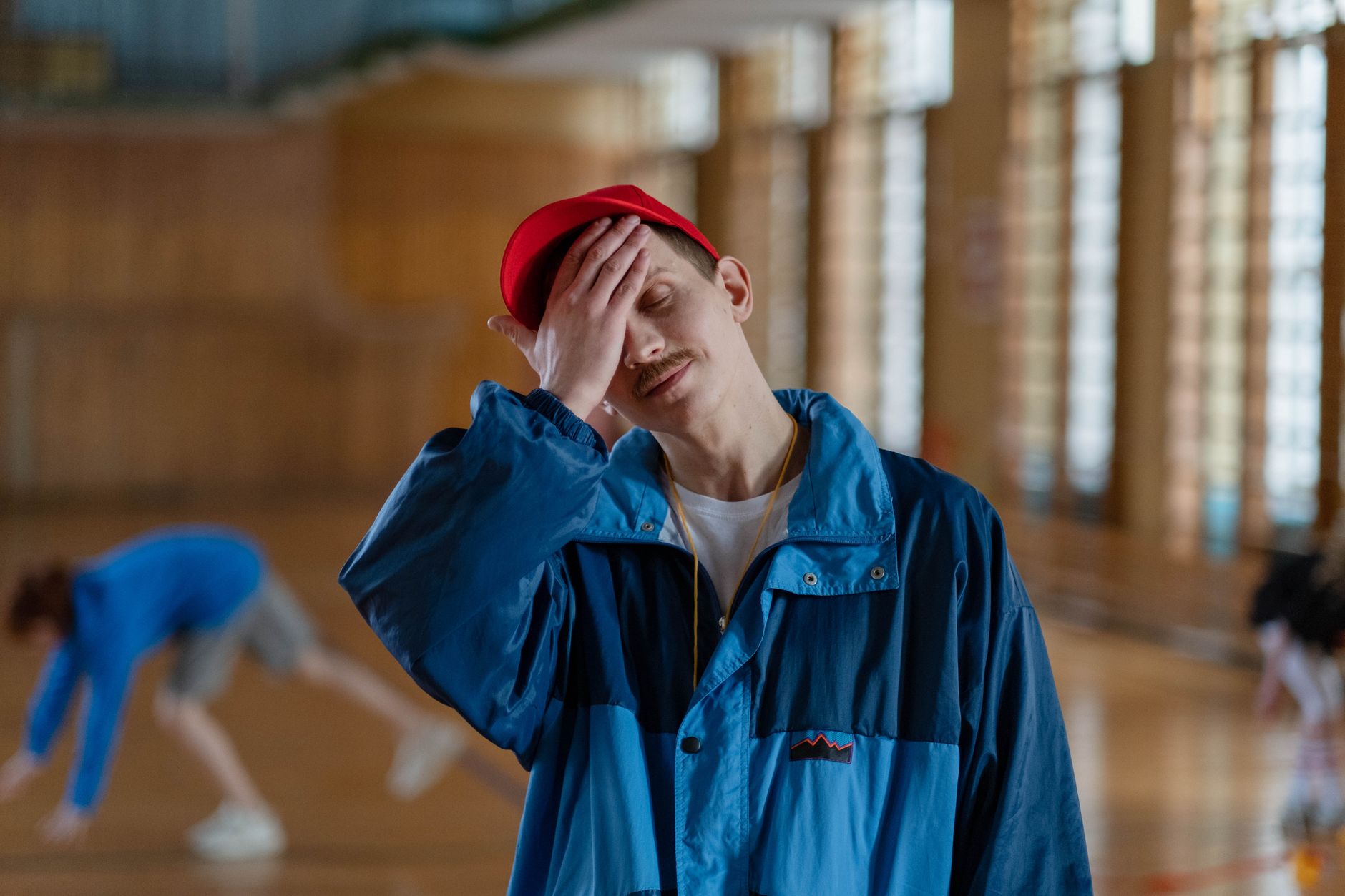A male physical educator in a gym looks exhausted, touching his forehead as students exercise in the background.