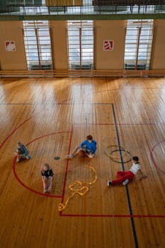 Kids are engaging in activities on a wooden sports hall floor with hula hoops and ropes.