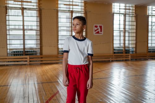 Free A young boy in sports attire stands confidently in an indoor gymnasium with wooden floors. Stock Photo