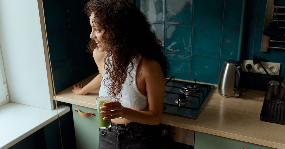 person looking energized and happy, holding a green smoothie, standing in a sunny kitchen
