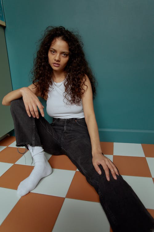 Woman in White Tank Top and Black Denim Jeans Sitting on Orange and White Floor Tiles