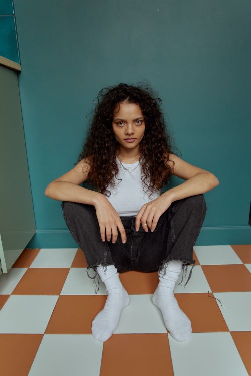 Free Portrait of a young woman with curly hair sitting on a tiled floor, exuding confidence. Stock Photo