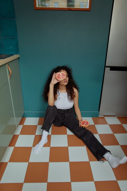 Free A young woman sits on a tiled floor, playfully holding grapefruits, indoors. Stock Photo
