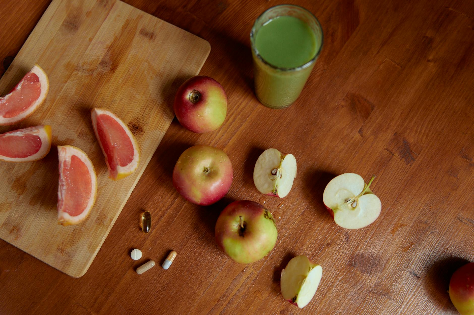 Top view of sliced grapefruit, apples, pills, and a green drink on a wooden table, emphasizing health.