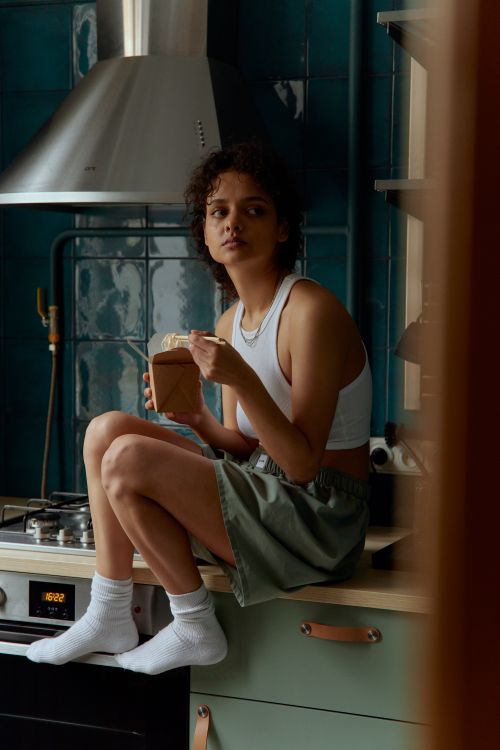Free A young woman in a casual outfit enjoys noodles while sitting on a kitchen counter. Stock Photo