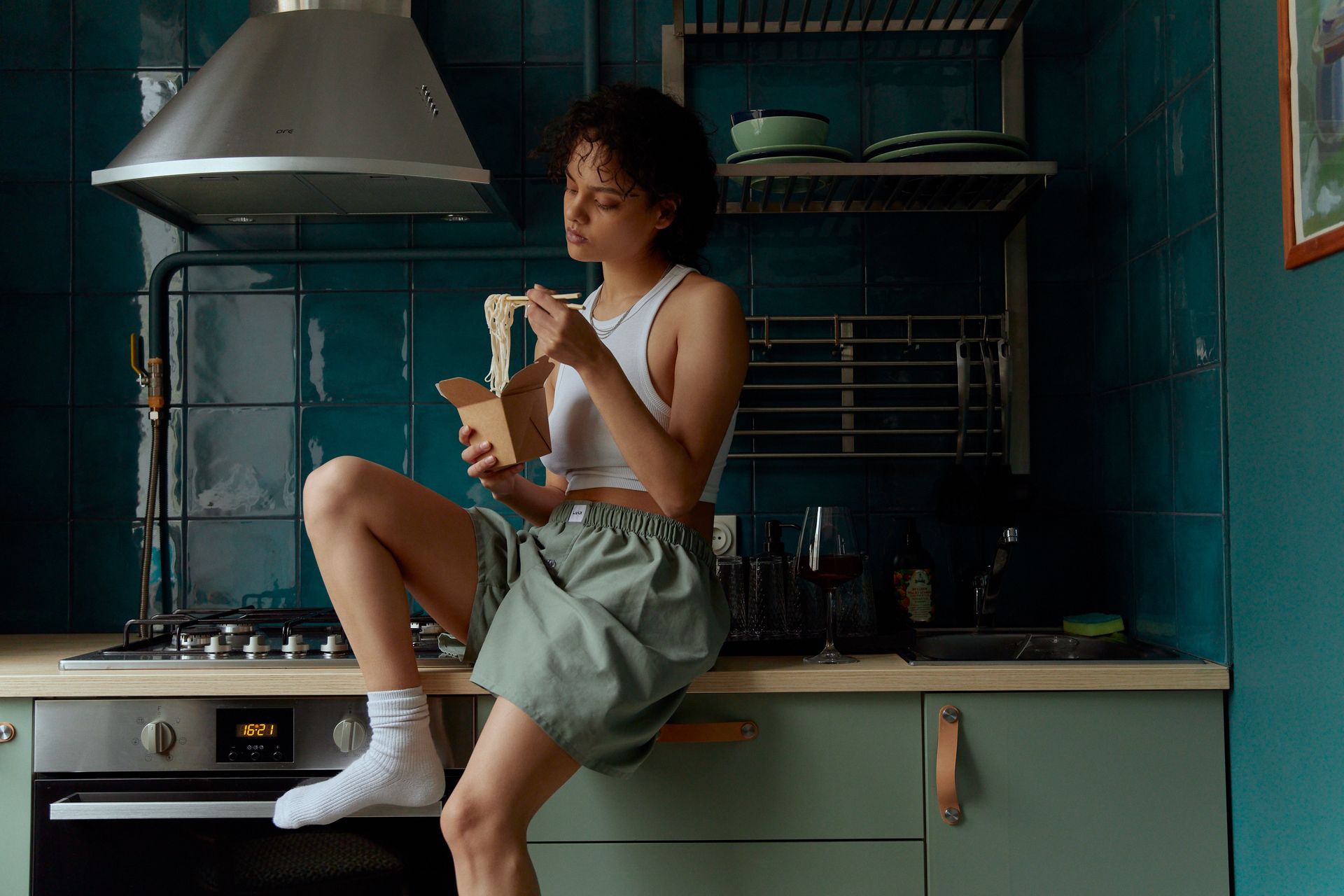 Young female in gray shorts and white top and socks eating noodles with sticks from carton box while sitting in kitchen in modern apartment