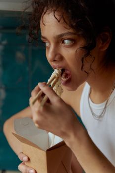 A young woman savoring a meal with chopsticks from a takeout box indoors.
