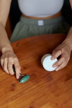 Close-up of hands using a jade roller and cream jar on a wooden table.