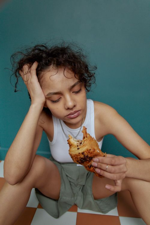 Free A young woman sitting on a tiled floor, thoughtfully eating bread indoors. Stock Photo