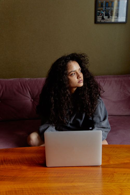 Free Pensive woman sitting indoors on a sofa with a laptop on a wooden table. Stock Photo