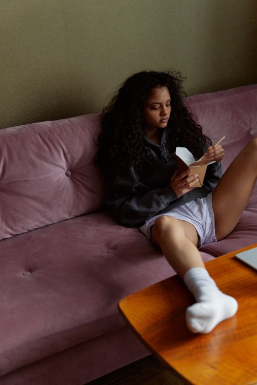 Free A woman sits on a pink sofa enjoying a meal from a takeout box in a cozy indoor setting. Stock Photo