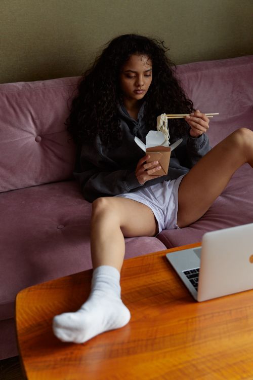 Free Woman sitting casually on a sofa, eating takeout noodles with a laptop nearby. Stock Photo