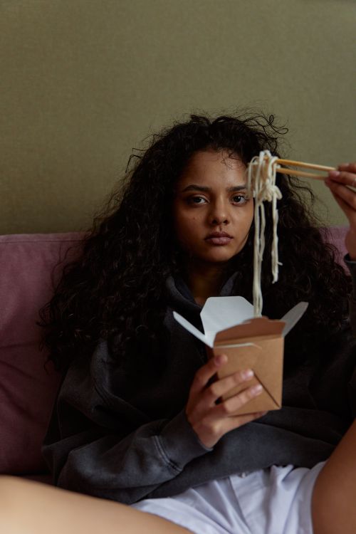 Free Young woman enjoying noodles from a takeout box while sitting indoors, appearing thoughtful. Stock Photo