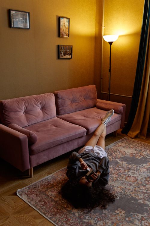 Free Woman in cozy living room lying on carpet, using phone, next to pink sofa with warm lighting. Stock Photo
