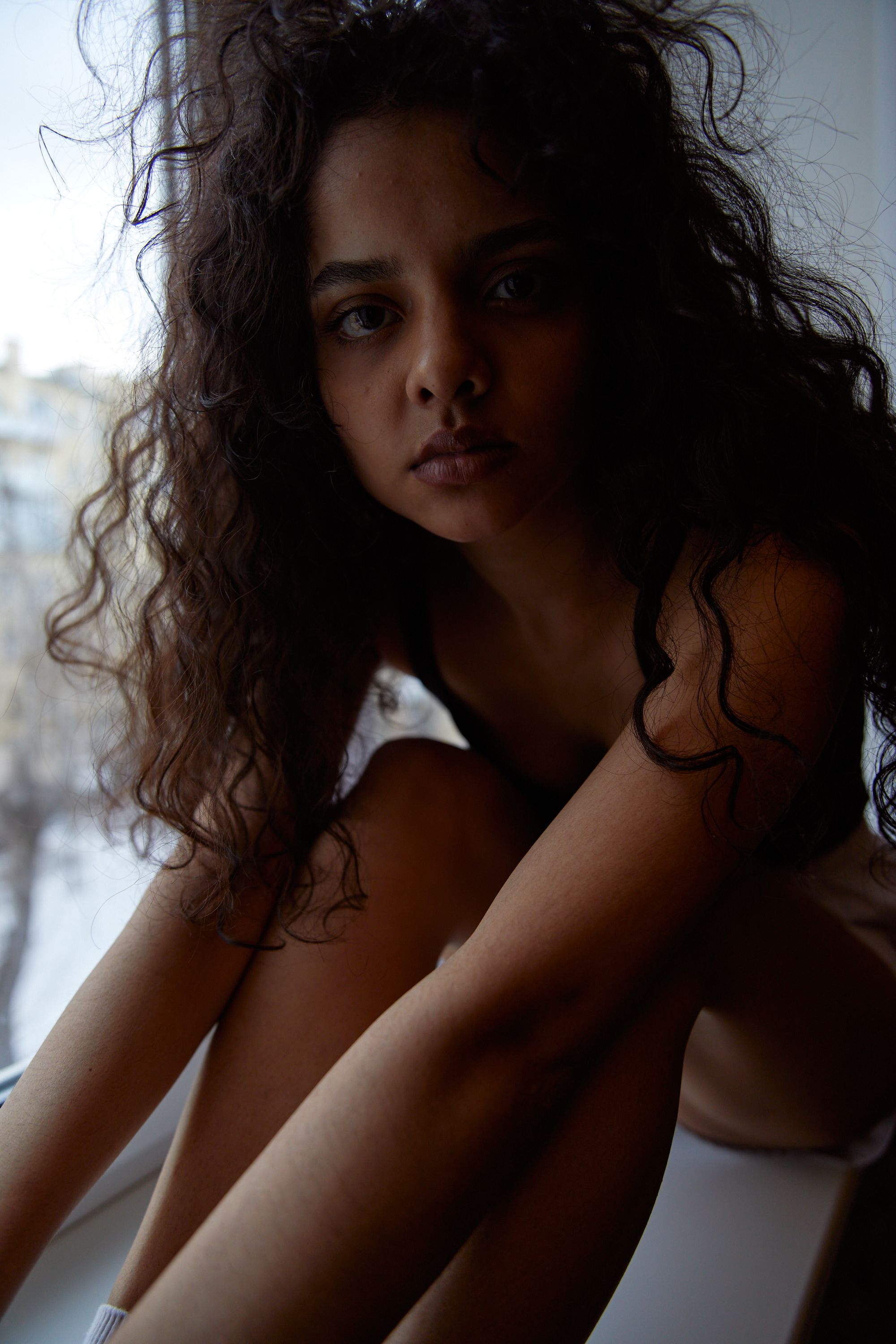 A woman with curly hair sits by a window, looking thoughtfully outside.