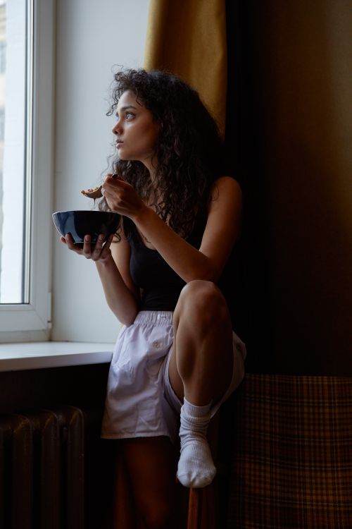 Free Woman sitting by the window, eating from a bowl, gazing thoughtfully outside. Stock Photo