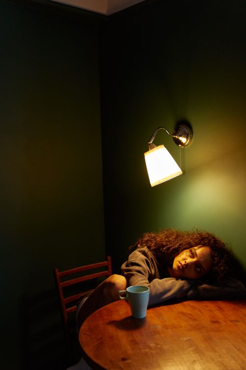 Free A woman with curly hair sits alone in a dimly lit room, expressing a contemplative mood. Stock Photo