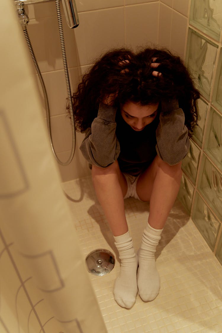 A Woman In Gray Sweater Sitting Inside The Bathroom With Her Hands On Her Head