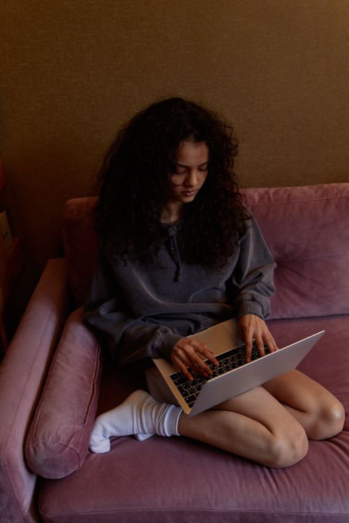 Free Young woman lounging on a pink sofa, typing on a laptop in a cozy indoor setting. Stock Photo