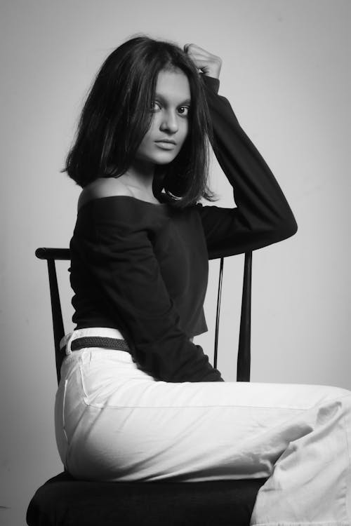 Black and white of young ethnic female in trendy outfit looking at camera on stool on white background of studio