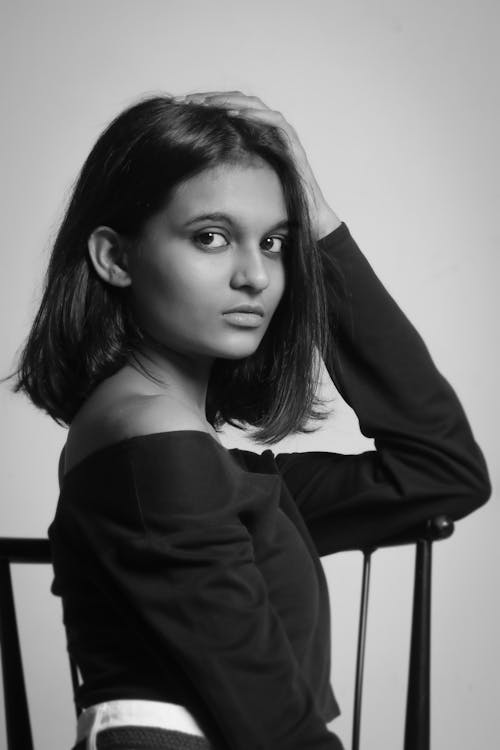 Black and white of young ethnic female with short hair on chair on white background of studio