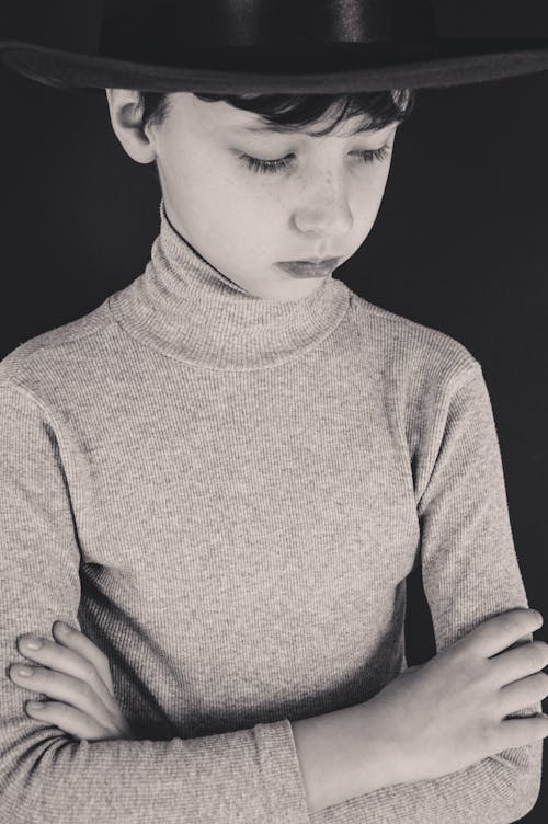 Black and white of serious boy in casual turtleneck and black hat looking down and holding arms crossed on black background