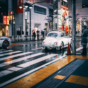 A rainy city intersection at night featuring a classic Volkswagen Beetle and modern storefronts.