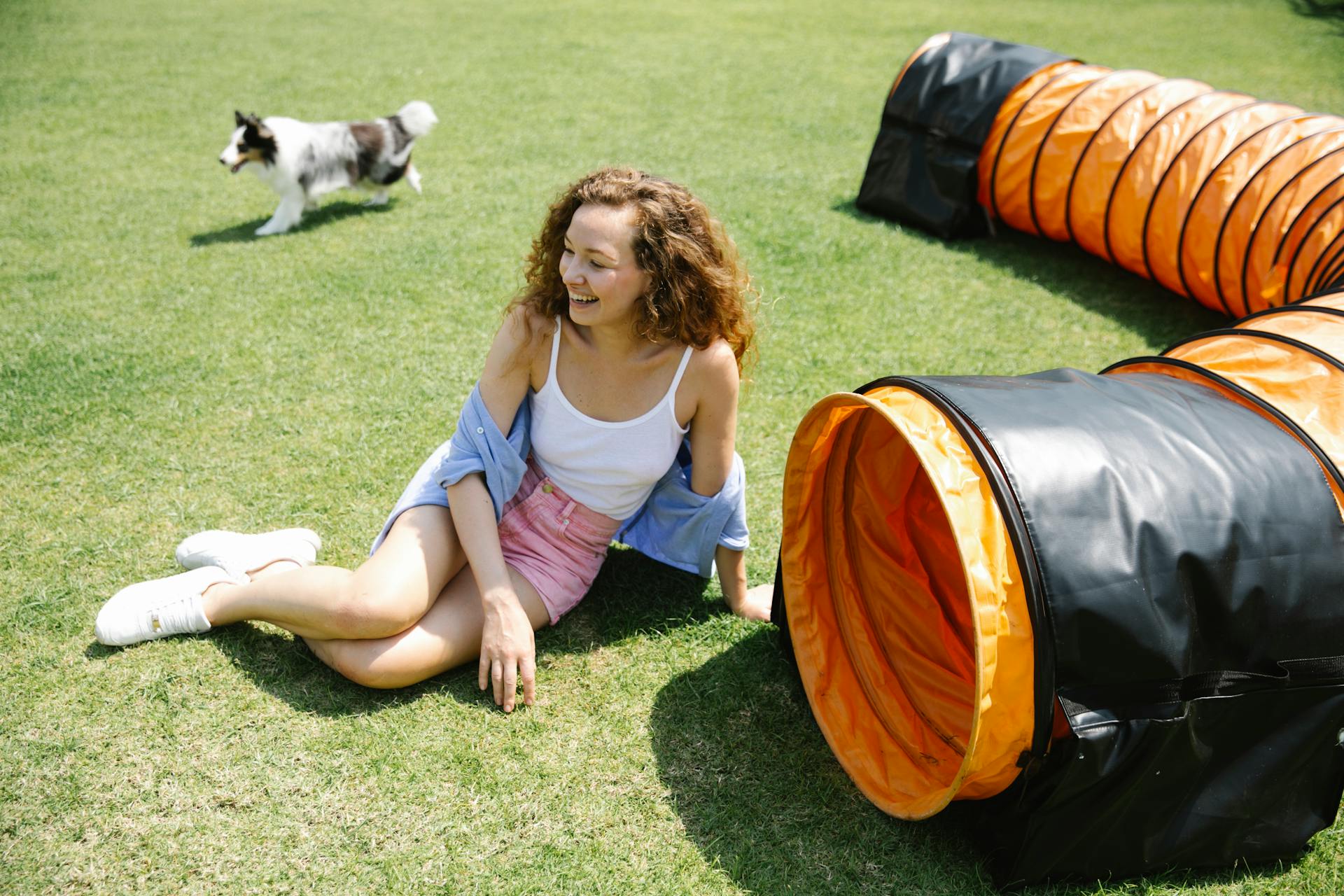 From above of laughing female with long curly hair sitting on green grass near playing tunnel while walking dog on sunny day in summer