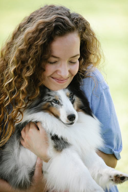 Smiling young female in casual clothes embracing fluffy dog on blurred background