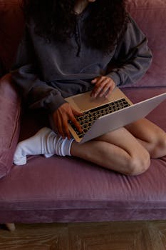 A young woman sits on a purple sofa using a laptop, showcasing a relaxed home environment.
