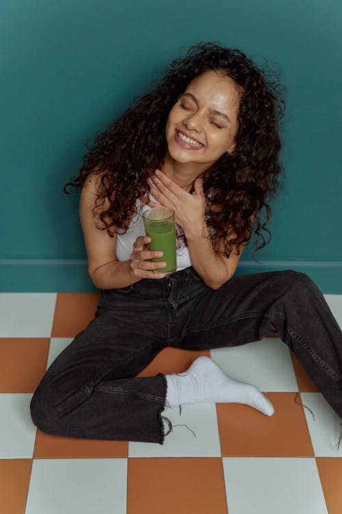 Woman in Black Tank Top and Blue Denim Jeans Holding Green Cup