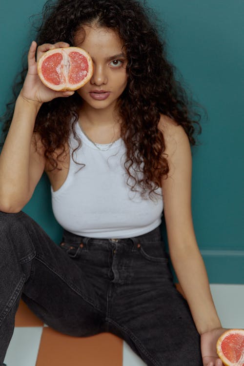 Free A Woman in White Tank Top Sitting while Holding a Sliced Grapefruit Stock Photo