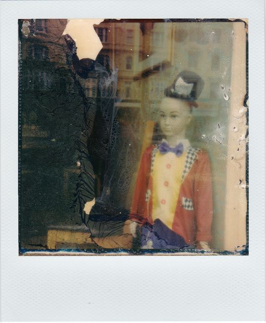 Retro Polaroid photo of a puppet in vibrant attire behind a glass window.