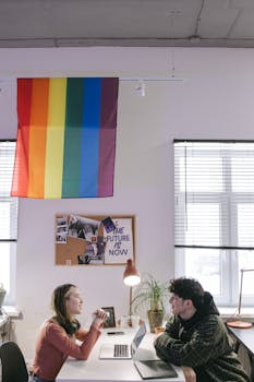 Two colleagues collaborate in a modern workspace with a pride flag displayed.