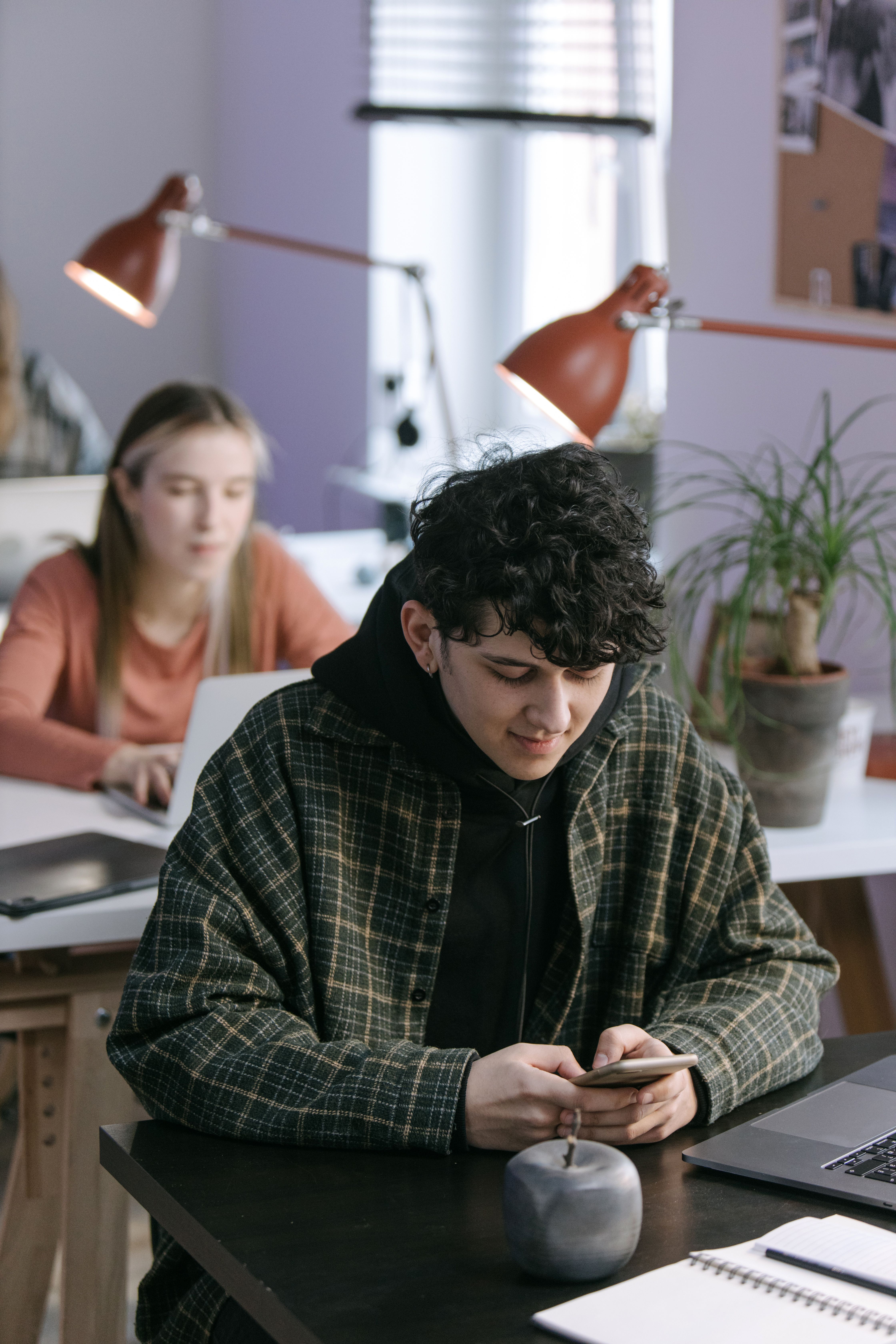 A young adult in a plaid jacket using a phone in a modern, well-lit workspace with laptops and plants.