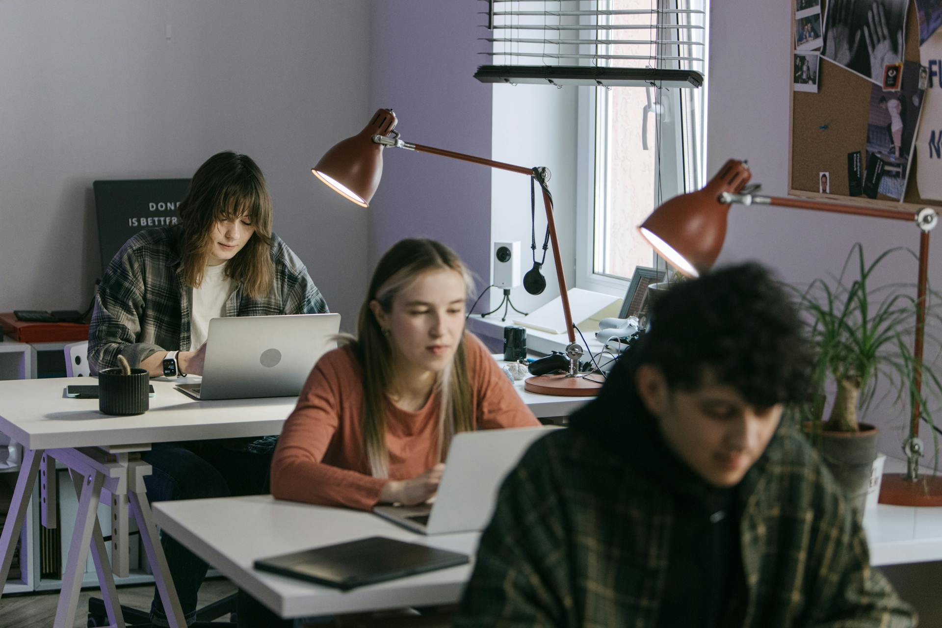 Three young professionals working at desks in a modern office setting with laptops and desk lamps.