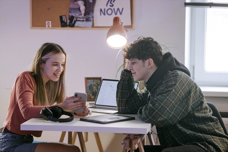 Young Man And Woman Having Conversation Sitting On A Table Using Smartphone