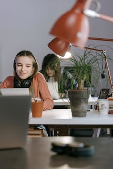 A young woman with headphones working on a laptop at a modern desk with plants and warm lighting.