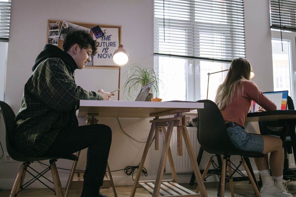 Dos jóvenes adultos concentrados en el trabajo en una oficina moderna, usando portátiles y teléfonos inteligentes.