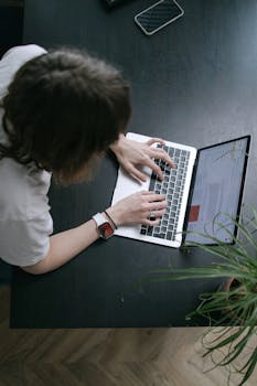 Overhead shot of a person typing on a laptop at a desk with a smartphone and plant in view.