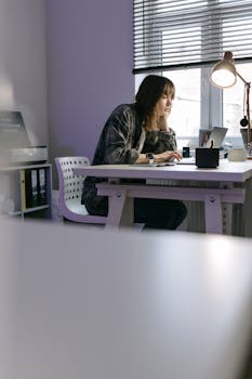 A woman working on a laptop in an indoor home office setting, focused on her task.