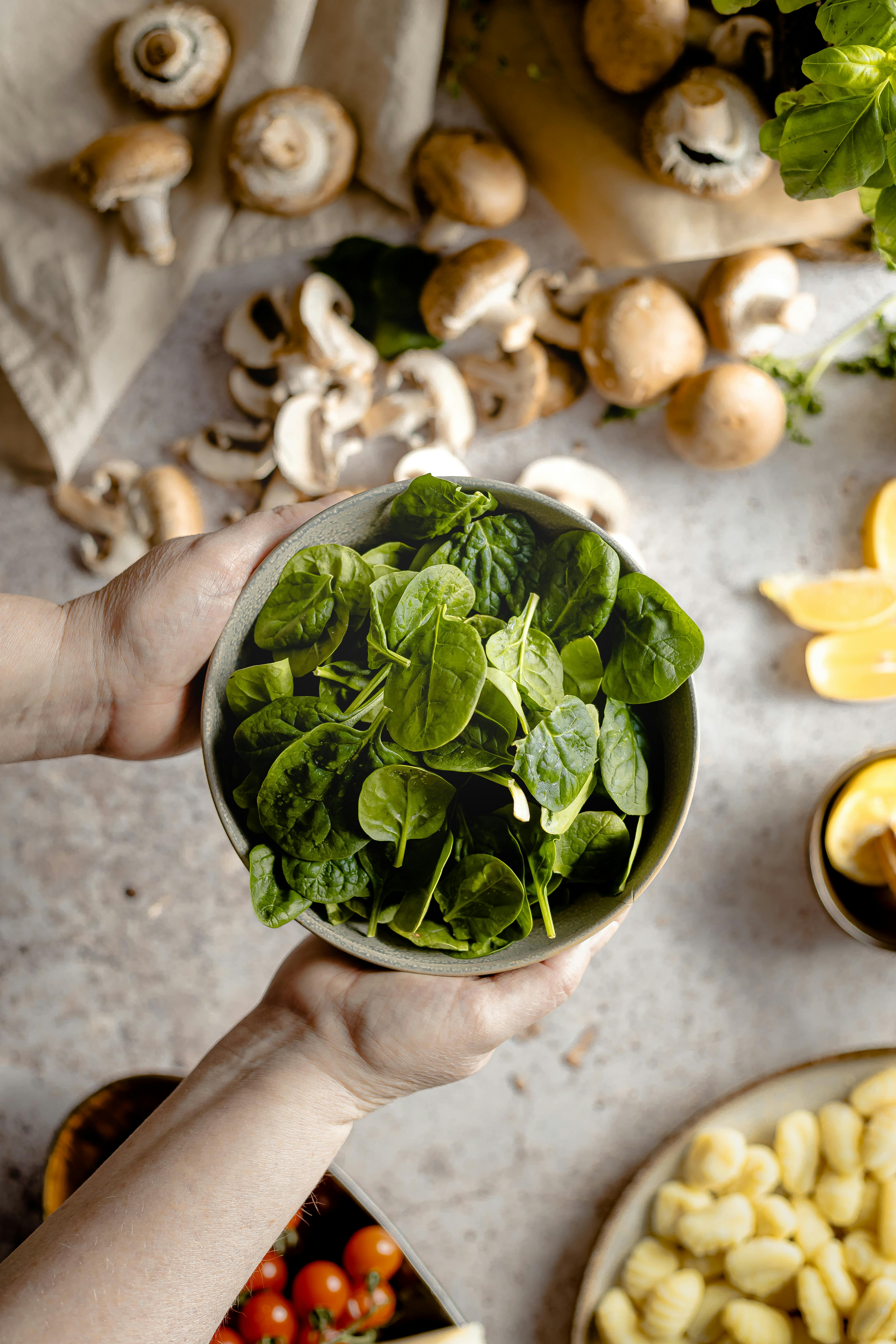 Top view of hands holding a bowl of fresh spinach with ingredients like mushrooms and tomatoes.