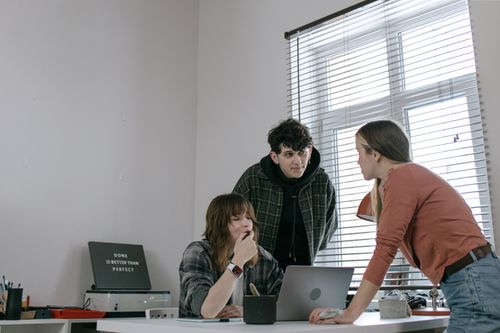 Free Team of young adults brainstorming ideas and discussing strategies around a laptop in a modern workplace. Stock Photo
