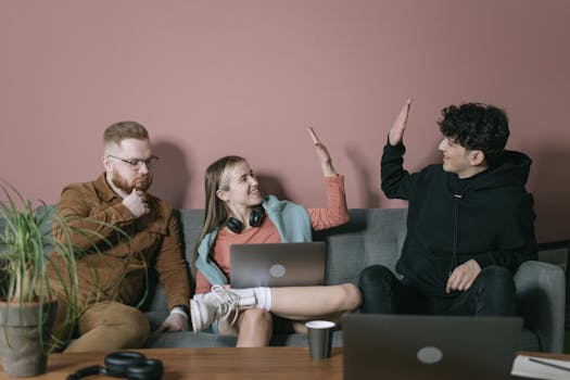 Three young adults collaborating indoors, sharing a fun moment with laptops and headphones.