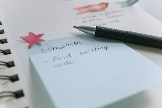 Close-up of a sticky note with a list, pen, and notebook on a desk setup.