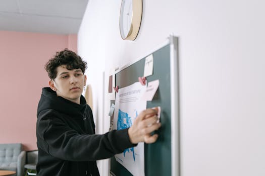 A young man in a hoodie interacts with a bulletin board, demonstrating collaboration in an office environment.