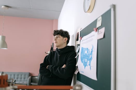 Young man in a modern office setting, leaning against a whiteboard with maps and notes, engaged in deep thought.