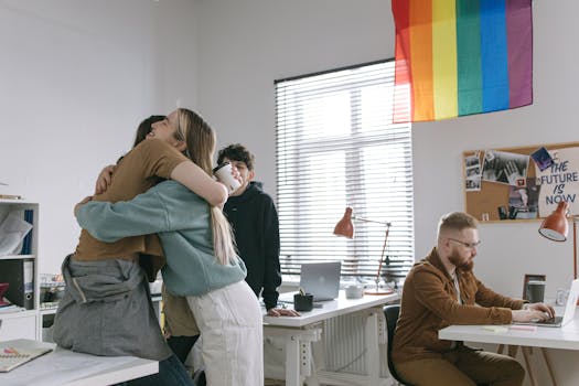 A group of diverse coworkers embrace and work on laptops under a rainbow flag.