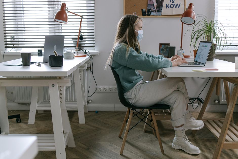 Young woman wearing mask, working on laptop at home office desk. Comfortable and modern workspace setup.