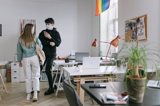 Two colleagues in masks interacting in a modern, light-filled office space with open workstations.